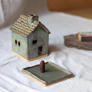 Puede incluir: Un porta incienso de cerámica con forma de casa, con un tejado texturizado y una pequeña chimenea. El soporte es de color verde apagado con una bandeja a juego y un cono de incienso marrón. La casa tiene dos ventanas y una puerta.