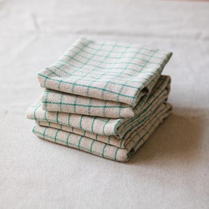 May include: A stack of five beige and green checked kitchen towels. The towels are folded and stacked neatly. The green grid pattern is visible on the top towel and the edges of the others. The towels are made of a natural fabric.