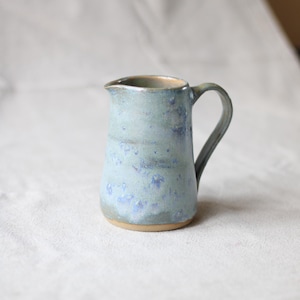 Può includere: Piccolo brocca in ceramica fatta a mano con smalto azzurro chiaro e motivo maculato. La brocca ha un manico curvo e un beccuccio per versare facilmente. La base è di colore argilla naturale non smaltata. Un articolo da cucina rustico e artigianale.