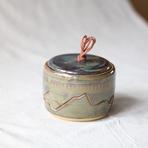 May include: A round ceramic jar with a lid featuring a glossy blue and green glaze. The exterior of the jar displays a carved mountain range pattern around the middle. A small tan leather cord is tied through the center of the lid as a handle.