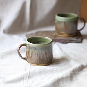 Könnte beinhalten: Zwei handgefertigte Keramikbecher mit rustikaler Ästhetik. Die Becher haben eine grüne und braune Glasur mit vertikalen, geätzten Linien. Ein Becher ist scharf, der andere ist leicht verschwommen im Hintergrund. Die Becher stehen auf einem weißen Tuch.