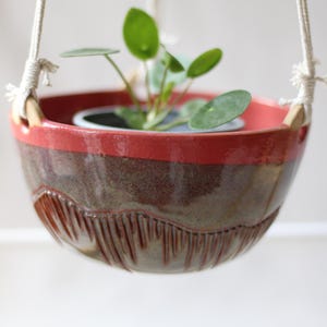 Può includere: Fioriera sospesa in ceramica con bordo rosso e corpo marrone strutturato. La fioriera è sospesa da una corda bianca e contiene una piccola pianta verde con foglie rotonde. Un oggetto decorativo e funzionale.