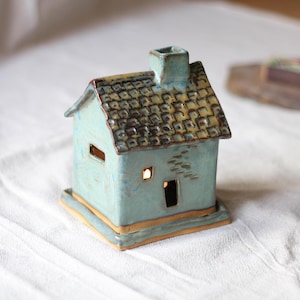 Può includere: Un portacandele in ceramica a forma di casa color turchese con un tetto di tegole marroni e gialle, un camino e finestre. Il portacandele poggia su una base quadrata con un bordo dorato. Le finestre brillano di luce.