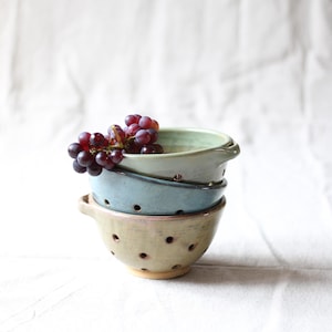 Könnte beinhalten: Ein Stapel aus drei Keramikschalen für Beeren mit kleinen Abflusslöchern und seitlichen Griffen. Die oberste Schale ist hellgrün, die mittlere blau und die unterste beige mit einem rosafarbenen Rand. Eine Traube dunkler lila Weintrauben liegt darin.