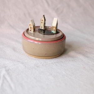 Puede incluir: Un tarro de cerámica redondo con tapa, acabado en color beige liso y borde rosa. La tapa presenta tres pequeñas esculturas verticales en forma de casa, en tonos beige y blanco, dispuestas alrededor de una mancha circular oscura y brillante.