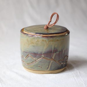 Peut inclure: Un pot cylindrique en céramique avec un couvercle muni d'une boucle de traction en cuir beige. L'extérieur présente une finition émaillée dans des tons sourds de vert sauge, de brun et de crème, avec un motif de montagne gravé sur la base.