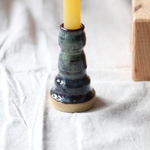 May include: A handcrafted ceramic candlestick with a yellow candle. The candlestick has a dark blue and green glaze with a beige base. The candle holder is approximately 13 cm tall.