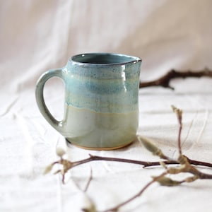 Può includere: Brocca in ceramica fatta a mano con manico arrotondato e bordo leggermente svasato. La brocca presenta una sfumatura di colori, che vanno da un verde-blu scuro nella parte superiore a un verde pallido alla base. Sfondo neutro.