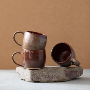 Può includere: Tre tazze in ceramica marrone con una glassa macchiata. Le tazze sono impilate su una roccia grigia.