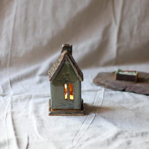 Peut inclure: Un photophore en céramique en forme de maison avec un toit texturé et une cheminée. La maison est de couleur vert clair avec une fenêtre rectangulaire éclairée d'une lumière chaude. La base et les bordures sont marron foncé. Une boîte d'allumettes est posée sur une dalle de pierre.