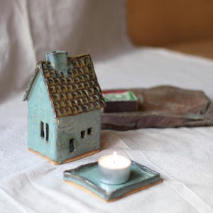 Può includere: Una casetta in ceramica turchese fatta a mano con tetto in tegole marroni e piccole finestre. Una candela turchese accesa è su un vassoio quadrato abbinato. La casa e il vassoio sono su una superficie bianca.