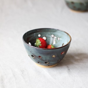 Op de afbeelding: Handgemaakte keramische kom met een teal glazuur en kleine gaatjes. De kom bevat verse rode aardbeien. De kom heeft een lichtbruine basis en een donkere rand. De achtergrond is een zachte, neutrale kleur.