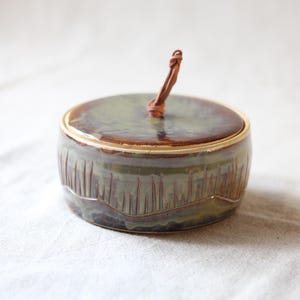 May include: A ceramic jar with a lid and a brown leather loop handle. The jar has a green and brown glaze with vertical etched lines around the base. The lid is a matching green and brown colour.