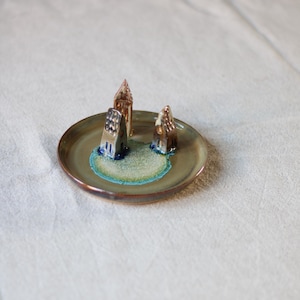 Puede incluir: Un plato decorativo de cerámica con tres casas en miniatura. Las casas son marrones, beige y azules, y están sobre una isla verde y azul. El plato es de un verde apagado con un borde color cobre.