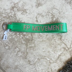 Pode incluir: Um porta-chaves verde brilhante com o texto "FP MOVEMENT" em strass prateados. O porta-chaves tem um anel de metal prateado e uma pequena etiqueta. Est&aacute; sobre um ch&atilde;o de ladrilhos de cor clara.