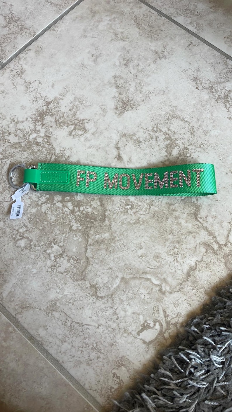 Pode incluir: Um porta-chaves verde brilhante com o texto "FP MOVEMENT" em strass prateados. O porta-chaves tem um anel de metal prateado e uma pequena etiqueta. Est&aacute; sobre um ch&atilde;o de ladrilhos de cor clara.