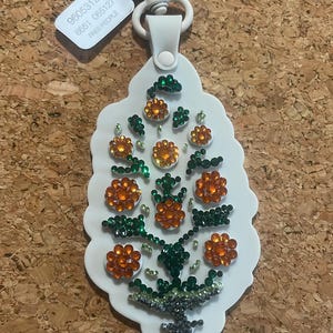 May include: White keychain with a floral design. The keychain features orange and green rhinestones arranged in a flower pattern. The keychain has a white strap and a silver-colored clasp. The tag reads "FREE PEOPLE".