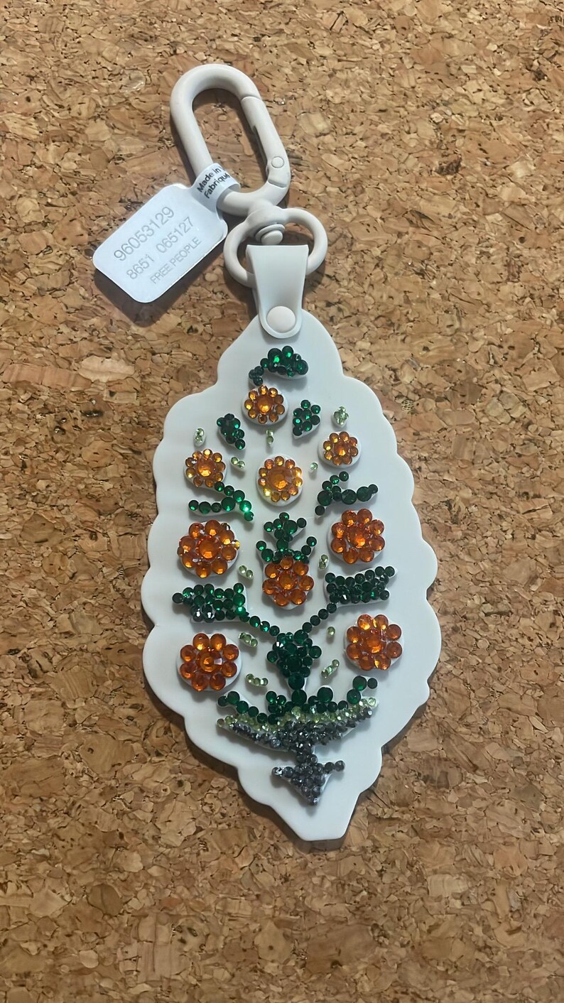 May include: White keychain with a floral design. The keychain features orange and green rhinestones arranged in a flower pattern. The keychain has a white strap and a silver-colored clasp. The tag reads "FREE PEOPLE".