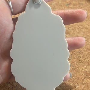 May include: A light gray, scalloped-edge ornament with a loop for hanging. The ornament is held in a hand, and a price tag is visible. The background is a corkboard surface.