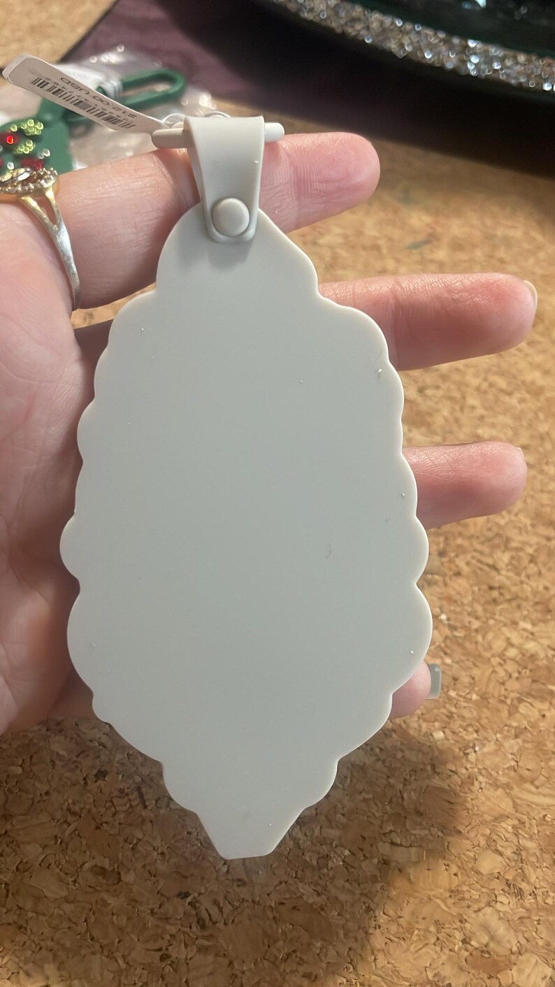 May include: A light gray, scalloped-edge ornament with a loop for hanging. The ornament is held in a hand, and a price tag is visible. The background is a corkboard surface.