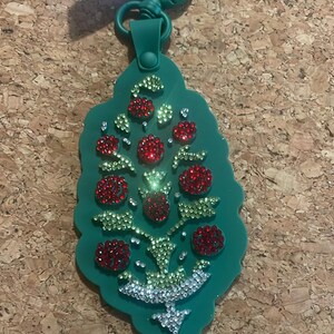 May include: A green keychain with a decorative tree design. The tree is adorned with red, green, and clear rhinestones. The keychain has a green clasp and a tag with the text "FREE PEOPLE".