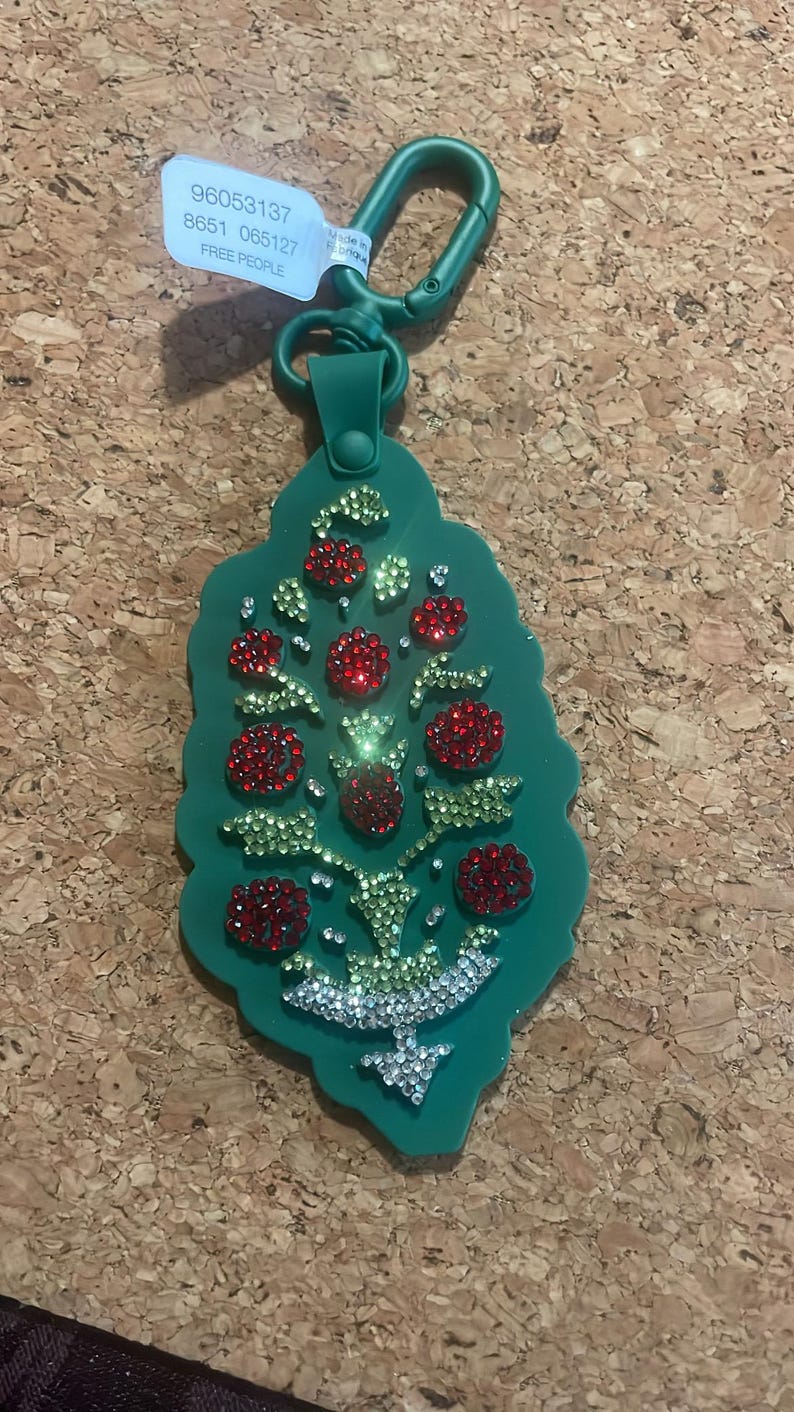 May include: A green keychain with a decorative tree design. The tree is adorned with red, green, and clear rhinestones. The keychain has a green clasp and a tag with the text "FREE PEOPLE".