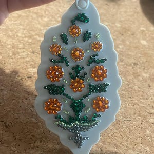 May include: A light gray decorative ornament with a floral design. The ornament features orange and green jewel-like embellishments arranged in a flower pattern. The text "FREE PEOPLE" is visible on the attached tag.