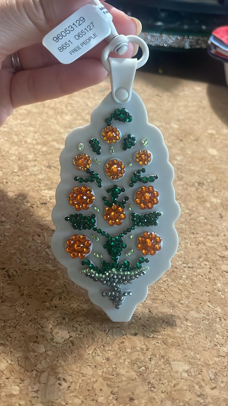 May include: A light gray decorative ornament with a floral design. The ornament features orange and green jewel-like embellishments arranged in a flower pattern. The text "FREE PEOPLE" is visible on the attached tag.