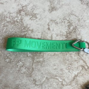 Pode incluir: Uma al&ccedil;a de chaveiro verde brilhante com o texto "FP MOVEMENT" na mesma cor verde. A al&ccedil;a tem um tri&acirc;ngulo e um anel de metal prateado para prender chaves. O item est&aacute; sobre uma superf&iacute;cie de azulejos de cor clara.