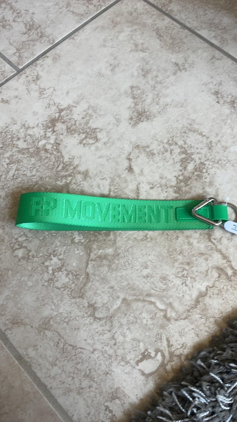 Pode incluir: Uma al&ccedil;a de chaveiro verde brilhante com o texto "FP MOVEMENT" na mesma cor verde. A al&ccedil;a tem um tri&acirc;ngulo e um anel de metal prateado para prender chaves. O item est&aacute; sobre uma superf&iacute;cie de azulejos de cor clara.
