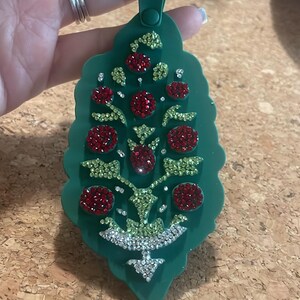May include: Green keychain with a leaf-like design, adorned with red and green rhinestones. The keychain has a green clasp and a tag with white text. The design features a tree-like pattern with sparkling accents.