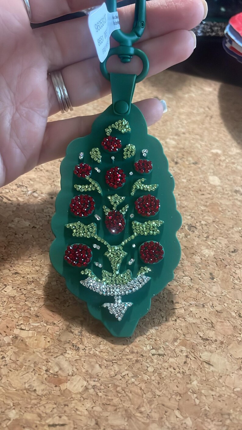 May include: Green keychain with a leaf-like design, adorned with red and green rhinestones. The keychain has a green clasp and a tag with white text. The design features a tree-like pattern with sparkling accents.