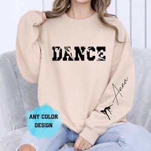 Puede incluir: Una sudadera beige con la palabra "DANCE" en letras negras, con siluetas de bailarines integradas en las letras. La manga presenta el nombre "Anna" en cursiva y una silueta de bailarina. También está presente el texto "ANY COLOR DESIGN".