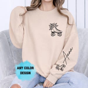 May include: A beige sweatshirt featuring a black roller skate design with floral details and the name "Anna" in script on the sleeve. The words "ANY COLOUR DESIGN" are in a blue watercolour splash.