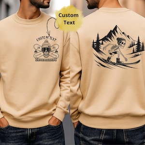 Könnte beinhalten: Beiges Sweatshirt mit Rundhalsausschnitt. Auf der Vorderseite befindet sich eine Grafik mit gekreuzten Skiern, einem Totenkopf und dem Text "Custom Text" und "Extreme Snowboarding 26". Auf der Rückseite ist ein snowboardendes Skelett zu sehen.