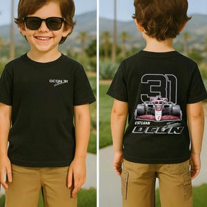 Op de afbeelding: Zwart T-shirt met de tekst "OCON 31" op de voorkant en een Formule 1 raceauto-afbeelding op de achterkant. De afbeelding bevat het nummer "31" en de naam "ESTEBAN OCON". Het shirt wordt gedragen met een kaki broek.