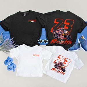Könnte beinhalten: Schwarze und weiße T-Shirts mit Motorradgrafiken und dem Namen "Bastianini". Ein Shirt zeigt die Nummer 23 in Rot. Ein Paar blaue Turnschuhe und eine Sonnenbrille sind ebenfalls im Bild. Die Shirts liegen auf einer blauen Oberfläche.