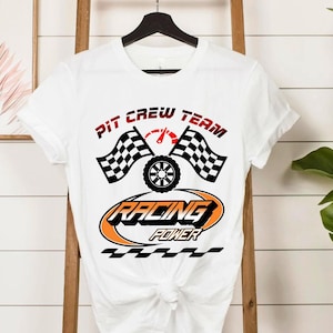 Puede incluir: Camiseta blanca con un diseño temático de carreras. El diseño incluye las palabras "PIT CREW TEAM" en rojo, dos banderas a cuadros, un neumático y las palabras "RACING POWER" en naranja y negro. La camiseta está colgada en una percha negra.