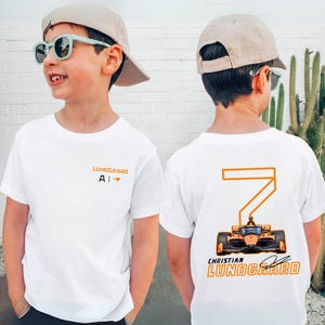 May include: White t-shirt with orange and black graphics. The front features the name "Lundgaard" and the letter "A". The back displays a large number "7" with a race car design and the name "Christian Lundgaard".