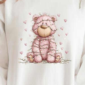 May include: White sweatshirt featuring a pink teddy bear wrapped in string lights. The bear has a light brown nose and is surrounded by pink hearts. The tag on the sweatshirt reads "WHITE".