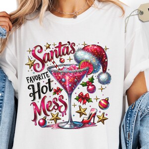 May include: White t-shirt featuring a festive design with a cocktail glass, Santa hat, and the text "Santa's Favorite Hot Mess." The design includes stars, ornaments, and a high heel, perfect for the holiday season.