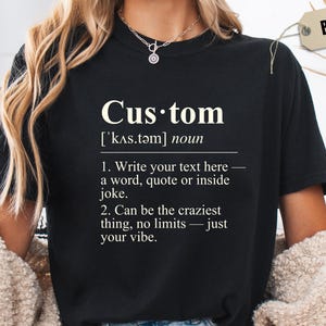 May include: Black t-shirt with the word "Custom" and its definition printed in white. The definition includes instructions to write your own text, quote, or joke. A tag on the shirt reads "BLACK".