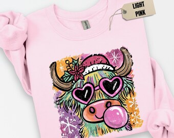 May include: Light pink sweatshirt featuring a colorful graphic of a Highland cow wearing heart-shaped sunglasses, a Santa hat, and blowing a bubble. The cow is surrounded by snowflakes and floral accents. The tag reads "LIGHT PINK".