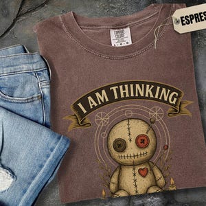May include: Espresso-colored t-shirt with the phrase "I AM THINKING" above a voodoo doll graphic. The doll has button eyes, a heart, and pins. A pair of blue jeans is folded next to the shirt.