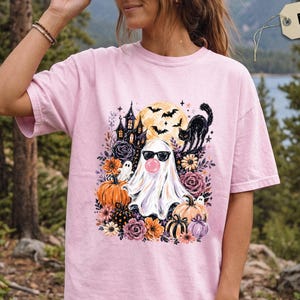 May include: A light pink t-shirt featuring a graphic print of a ghost wearing sunglasses and blowing a bubble, surrounded by a haunted house, a black cat, bats, a full moon, pumpkins, and various colourful flowers in shades of orange and purple.