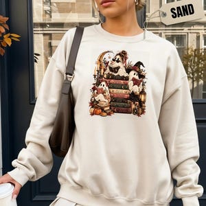May include: A sand-colored crewneck sweatshirt featuring a graphic print of three ghosts reading books, surrounded by pumpkins, candles, and a crescent moon. The design includes a stack of books and autumn foliage. A tag labeled SAND is visible.