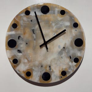 May include: A circular wall clock featuring a glossy, abstract background of blended gold, white, and grey tones. The clock face is accented with twelve solid black circular markers at each hour position and two simple black hands at the centre.