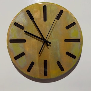 May include: A circular wall clock with a mottled yellow and gold finish. The clock face features twelve rectangular cutouts serving as hour markers and three thin black hands positioned at the centre to indicate the time on the dial.