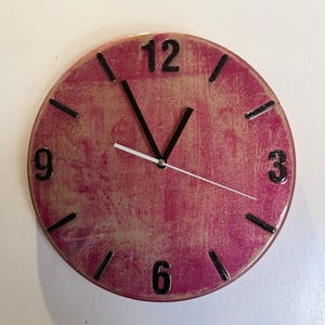 May include: A round wall clock with a textured magenta and gold metallic finish. The clock face features black numerals at the 12, 3, 6, and 9 o'clock positions, with black tick marks for the remaining hours. It has two black hands and a thin white second hand.