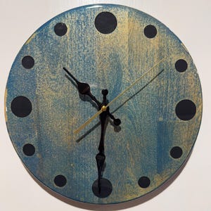 May include: A circular wall clock featuring a textured blue and gold painted face with twelve black circular hour markers. The clock has two black ornate hands and a thin gold second hand centred on the face, displaying a time of approximately 6:25.